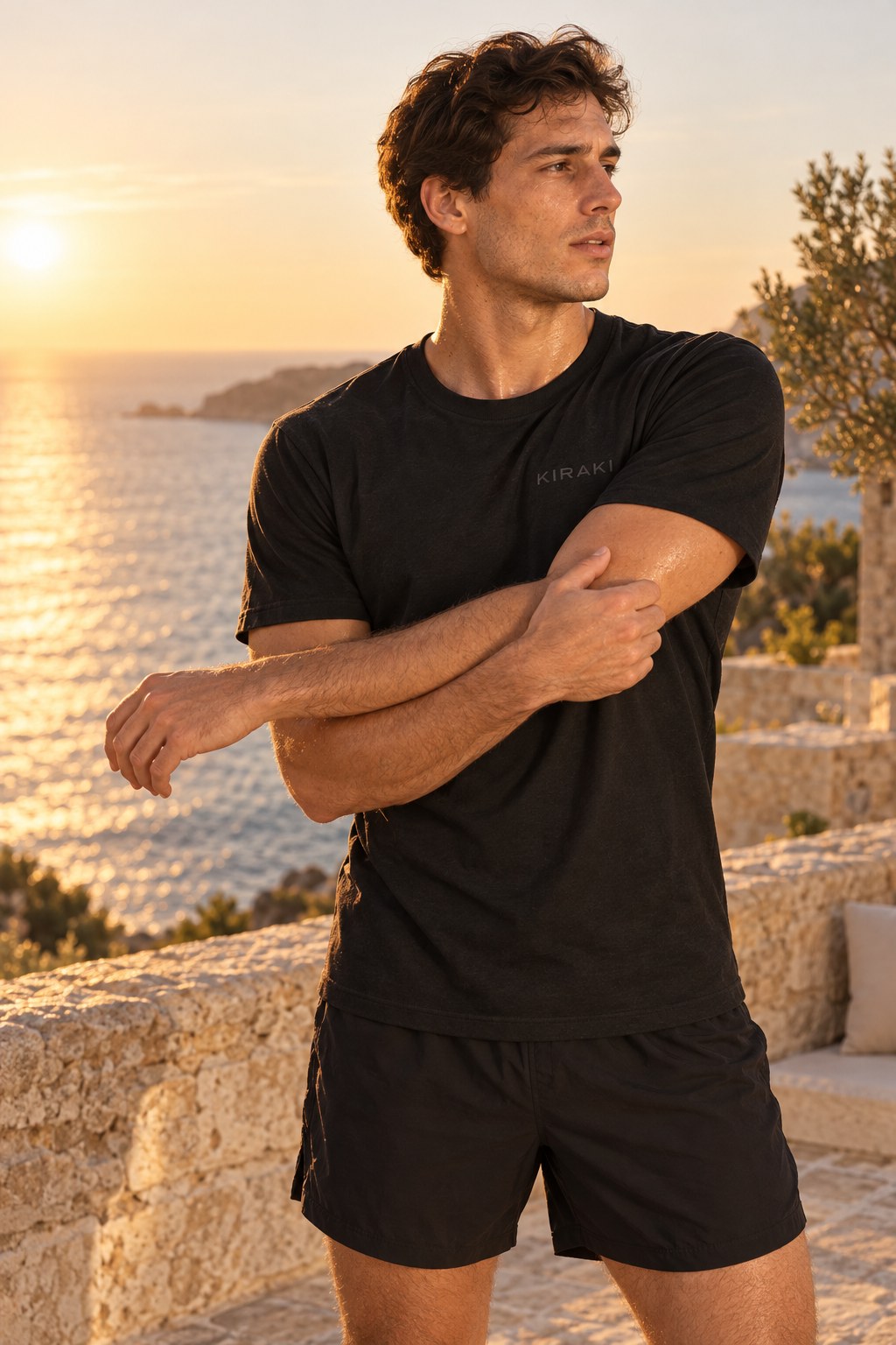 Man stretching on a sunlit stone terrace above the sea at golden hour, wearing the black KIRAKI Performance 01 tee