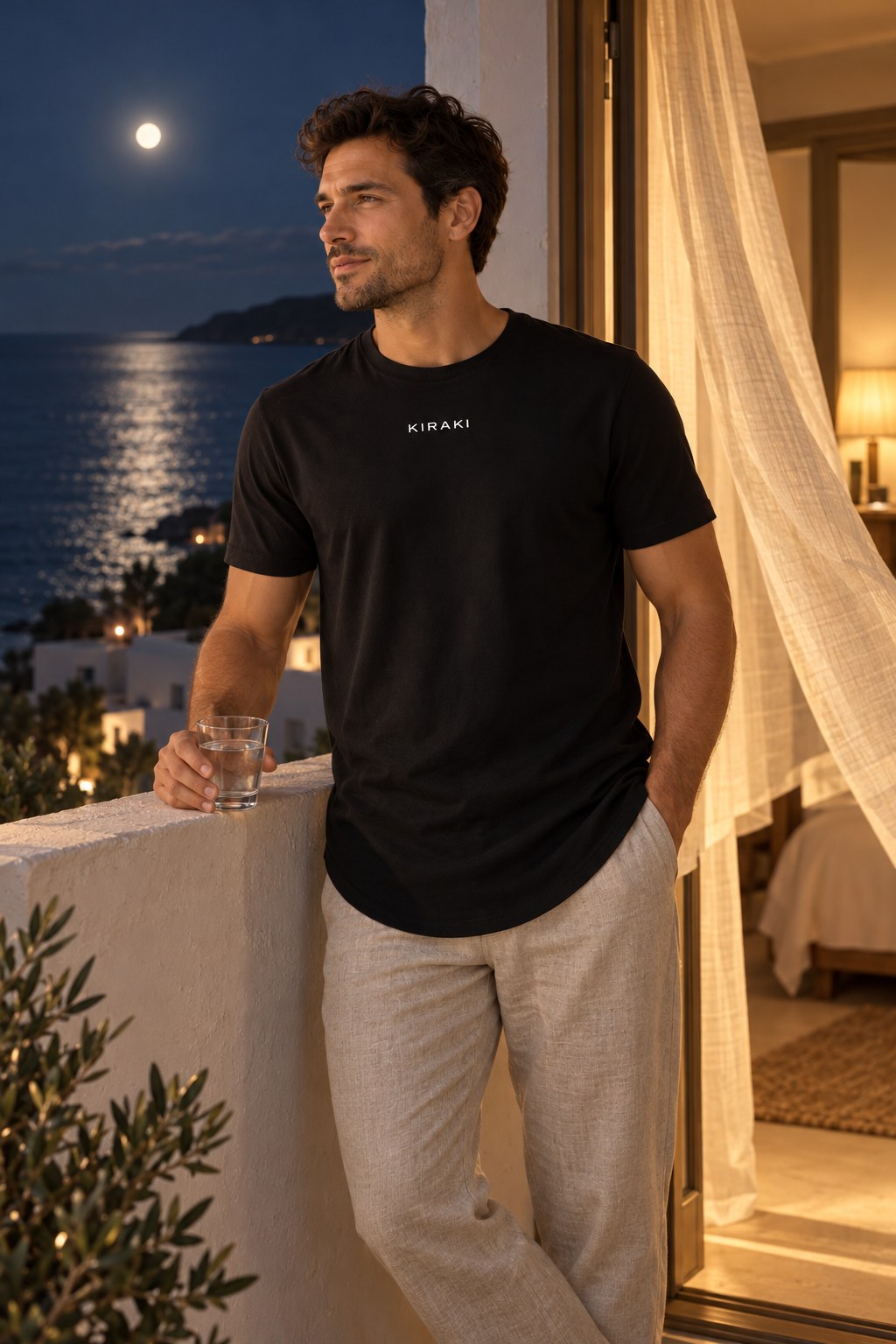 Man on a whitewashed balcony at night with a glass of water, moonlit sea behind, wearing the black KIRAKI Sleep 01 long-body tee